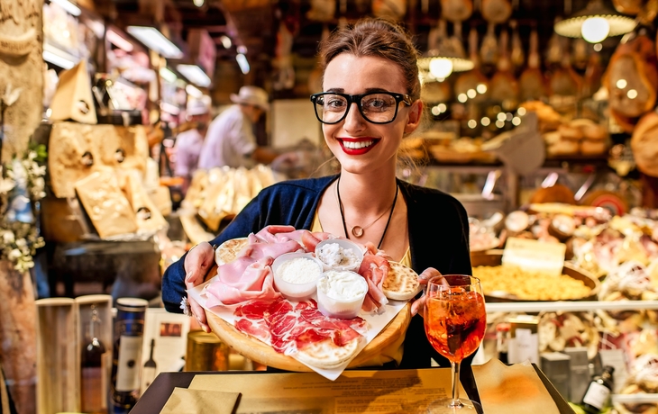 Junge Frau, die nahe dem Lebensmittelladen mit traditionellem italienischem Aperitif auf dem unscharfen Lebensmittelschaukastenhintergrund in der Bolognastadt sitzt. Bologna ist das gastronomische Zentrum Italiens